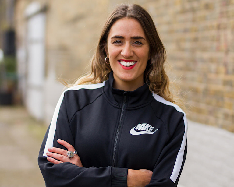 A headshot of Kelly Anna, against a brick wall. Kelly is a White woman in her thirties with brown hair, wearing red lipstick and a Nike sports jacket.