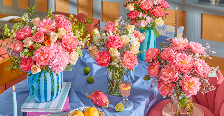 Bright kitchen scene with colorful floral bouquets (peonies, roses, more) in various vases on a light blue island. Champagne glasses, rosé, fruit, and decor add to the cheerful atmosphere.