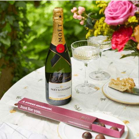 Bottle of Moët & Chandon champagne with glasses, pink roses, and a box of "A Sweet Moment" chocolates on an outdoor table.