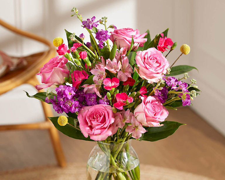 A clear vase holding a joyful bouquet of velvety pink roses, fragrant purple stocks and yellow craspedia. Lush greenery and hot pink blooms add texture. The bright, airy setting suggests a fresh, sweet summer fragrance.