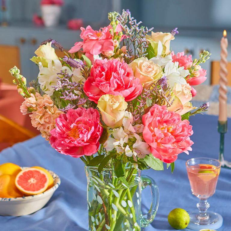 Bright yellow vase with pink peonies, purple and white wildflowers. Pink drink in a stemmed glass and yellow candle on a white counter. Teal cabinets in the background.