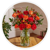 A bouquet of red and orange roses with winter greenery in a clear glass vase.