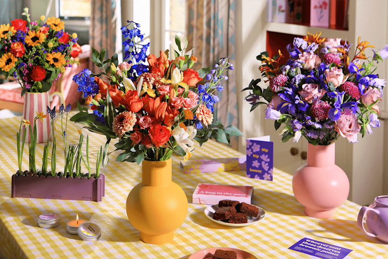 Drei bunte Blumensträuße in Vasen auf einem gedeckten Tisch mit Brownies, Kerzen und einer Grußkarte.