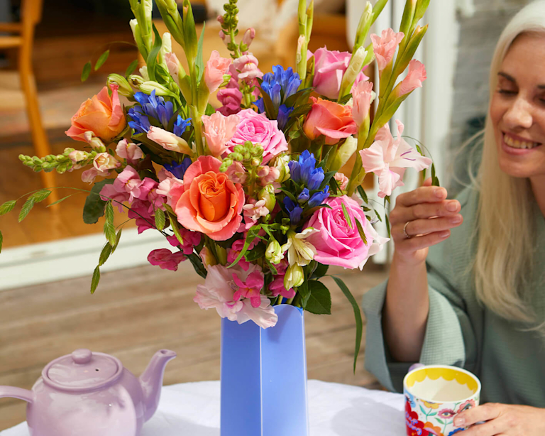 A woman with white hair admires a bouquet in a vase, a cup of tea in her hand. The bouquet is made up of bright, vibrant roses and gladioli.