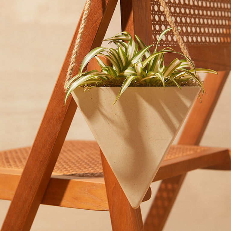 A spider plant in a beige, triangle-shaped planter hangs from a dark brown, wooden chair.