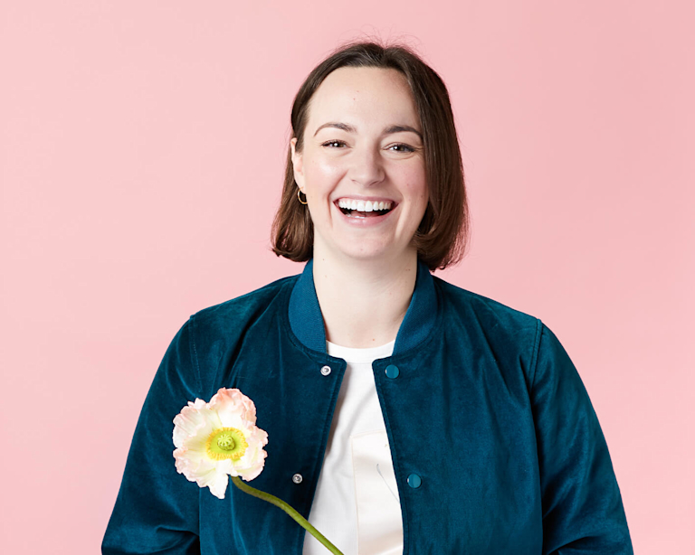 A White woman in her thirties with a short, brown bob smiles broadly at the camera. She wears a velvet bomber jacket and is holding a pink flower.