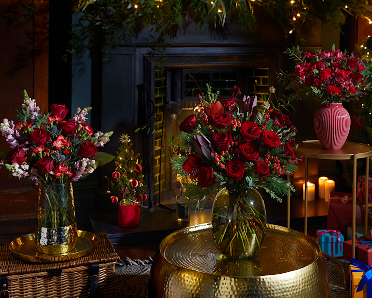Ein gemütliches Wohnzimmer mit einer Reihe an weihnachtlichen Blumensträußen in festlichen Farben. Im Hintergrund sind Geschenke und ein Weihnachtsbäumchen zu sehen.
