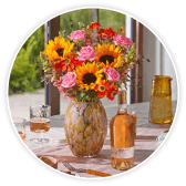 Sunflower and rose bouquet in a speckled glass vase on a table set with wine, glasses, and a striped runner by a window.