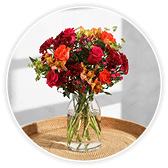 Vase of deep red, orange and burgundy roses and autumnal flowers in a clear glass vase, set on a woven rattan surface.