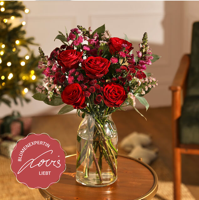 Romantischer Strauß mit dunkelroten Rosen und zarten pinken Beifüller-Blumen, arrangiert in einer Glasvase auf einem Beistelltisch vor festlicher Beleuchtung.