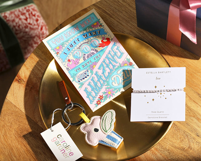 A flat-lay arrangement of gifts on a gold tray, including a colorful greeting card, a pink Rose Quartz gemstone bracelet by Estella Bartlett, and a cupcake-shaped leather keychain by Caroline Gardner.