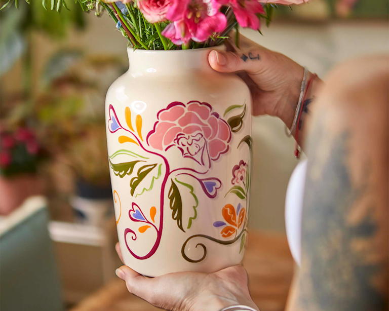 A person with tattooed arms holds a cream ceramic vase designed by Liv & Dom, filled with bright pink alstroemeria. The vase features a hand-painted, whimsical floral design in pink, orange, and purple. The scene feels artistic, tactile, and warm.