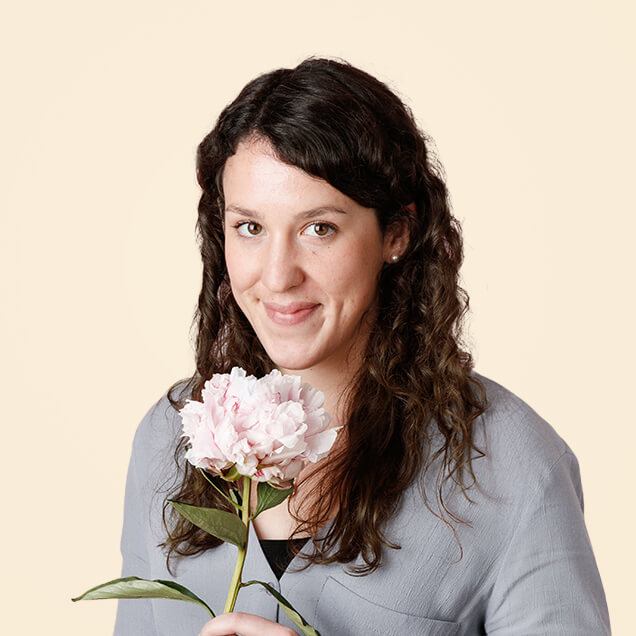 Lydia s a white woman in her thirties, with long dark curly hair. She holds a peony.