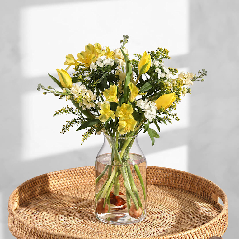 A sunny, uplifting arrangement of lemon-yellow tulips, buttery alstroemeria and ivory stocks. Presented in a clear vase on a woven tray.