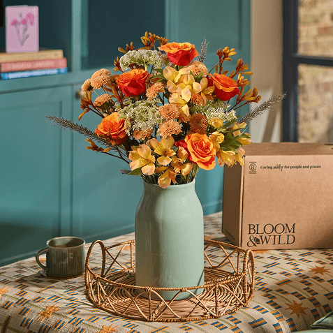 Orange roses, yellow alstroemeria, peach blooms, white ammi and dried grasses in a sage green vase on a woven rattan tray, beside a Bloom & Wild box and a green mug.
