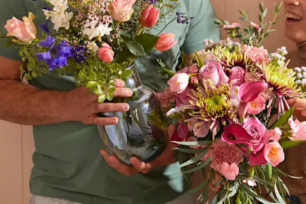 Two people are stood, holding abundant vases of vividly-coloured flowers.