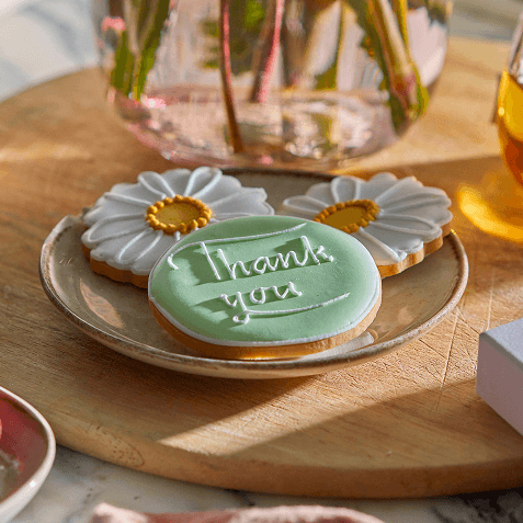 Daisy-shaped iced biscuits with a "Thank You" message, arranged on a rustic plate beside a pink glass vase in the sun.