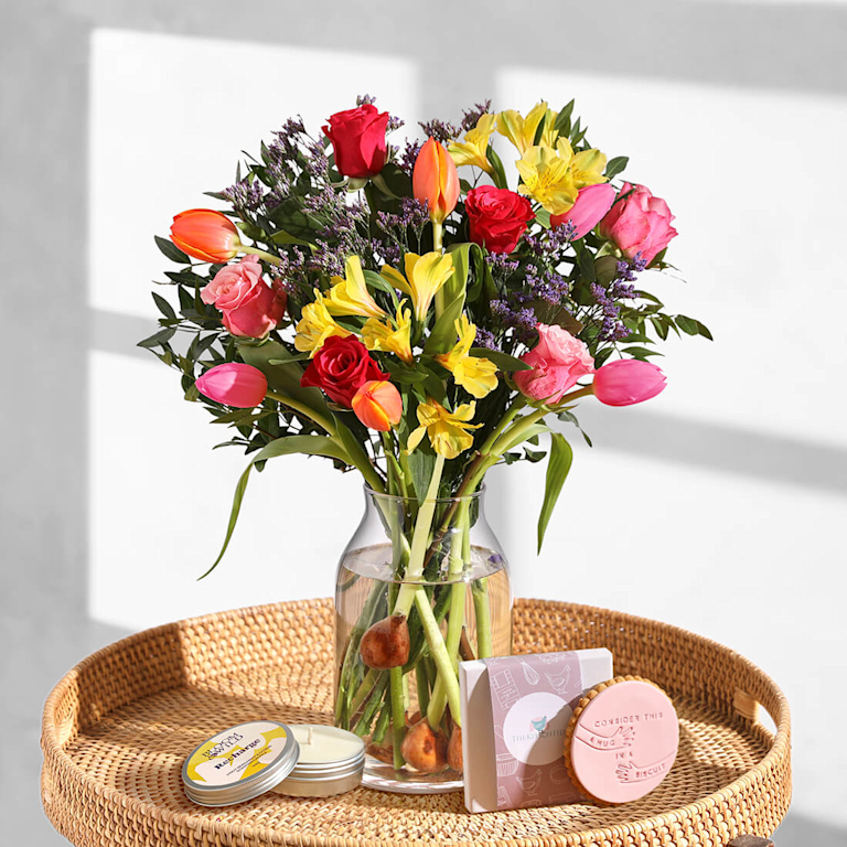 Bloom & Wild 'The Scottie' Bouquet featuring a mix of pink, orange and yellow tulips sits in a glass vase, alongside a small candle and iced biscuit.