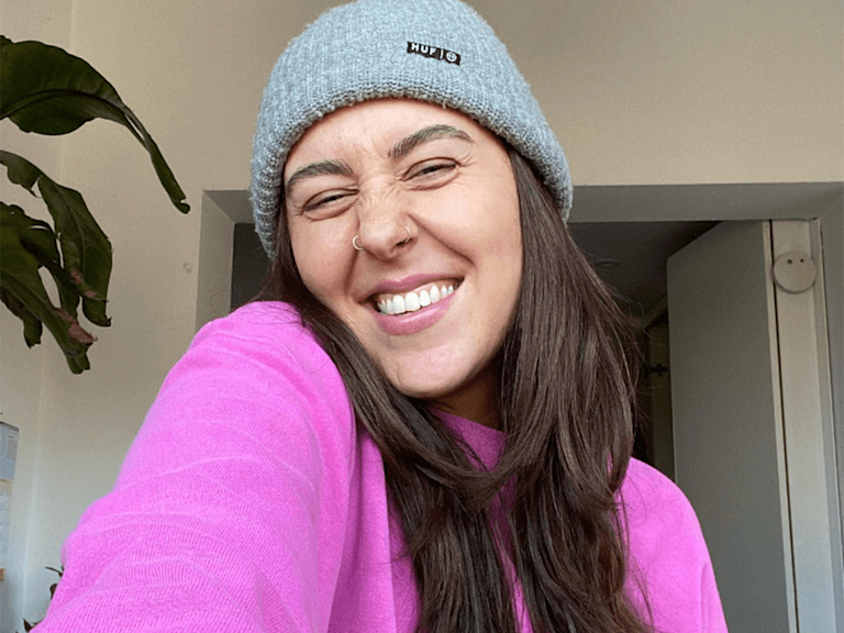 Gina, a light-skinned woman with nose-piercings, smiles playfully the camera. She wears a beanie and a hot pink jumper.