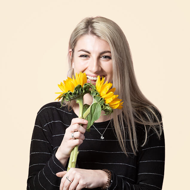 Jess is a white woman in her late twenties, with long blonde hair. She smiles as she holds two sunflowers over her face.