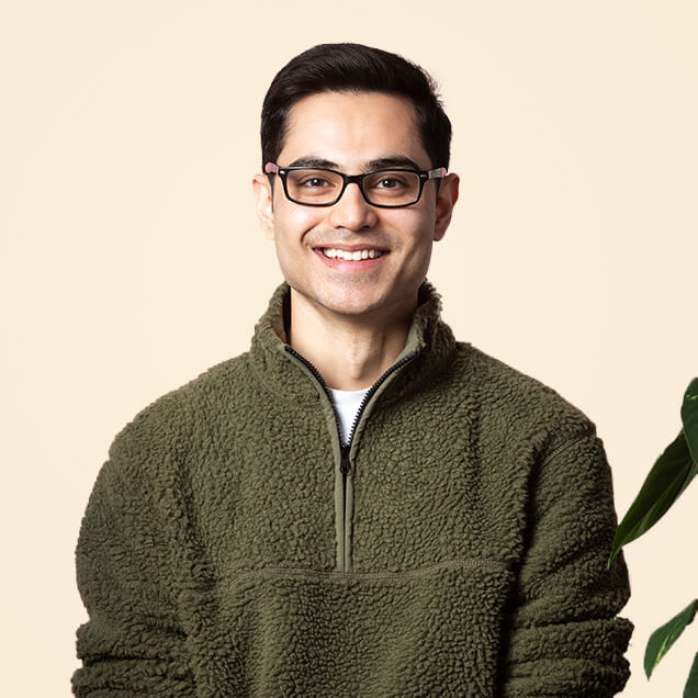 Syed is a man with a light-brown skin tone, dark hair and black glasses. He smiles next to a potted plant.