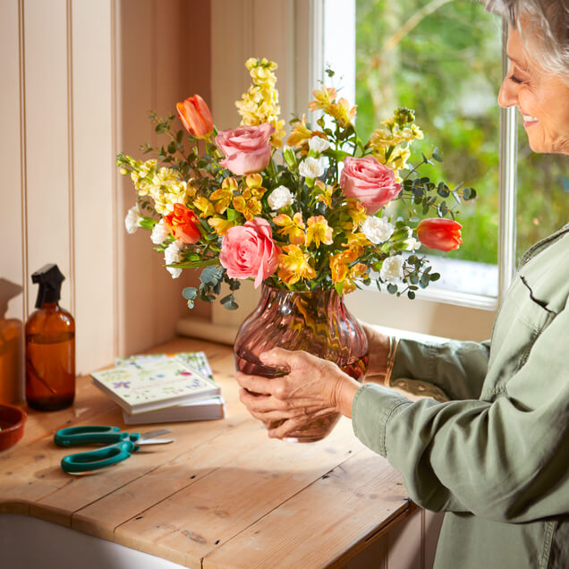 A split image shows a bright bouquet in a green and white checkered vase on a wicker table, with a "Happy Mother's Day" card and yarn balls beside it. The right side is a close-up of the bouquet, highlighting orange tulips, white irises, and purple tulips.