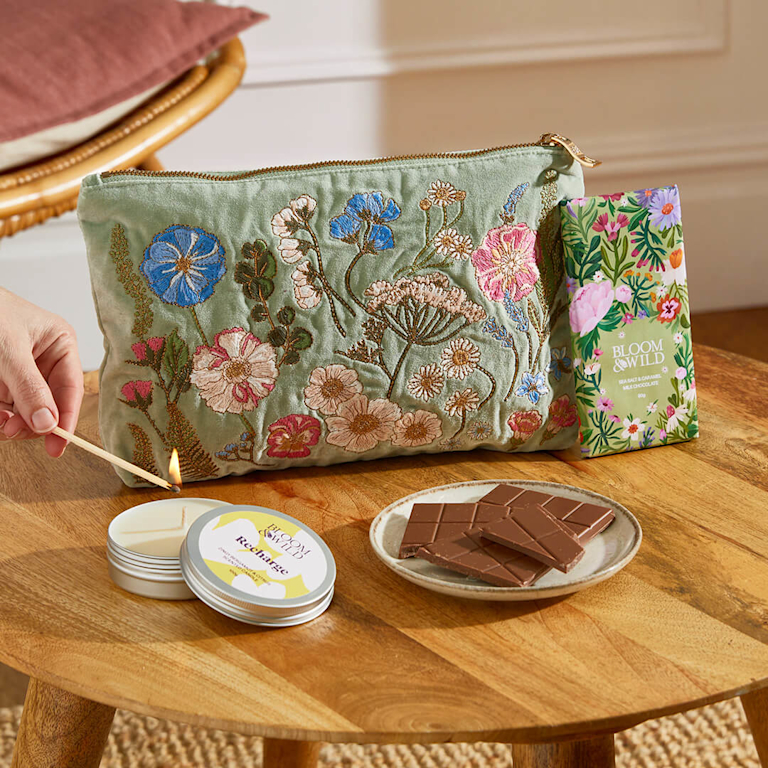 A self-care gift set featuring an intricate, floral-embroidered velvet pouch in sage green, a "Recharge" scented candle, and a bar of sea salt and caramel milk chocolate.