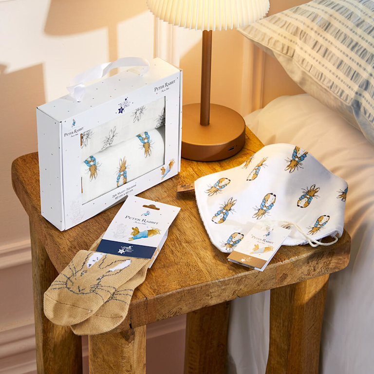 A charming Peter Rabbit-themed baby bundle, featuring a gift box of patterned muslin squares and sweet tan-coloured socks with bunny faces, all resting on a rustic wooden bedside table beside a lit lamp.