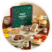 A green Happy Birthday treat box beside scones, cakes and sweet treats on a table.