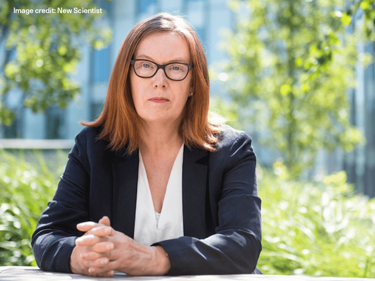 Sarah, a light-skinned older woman with glasses, wears a white shirt and blazer and sits on an outdoor table. Her hands are clasped in front of her, and she has a serious, quizzical expression on her face.
