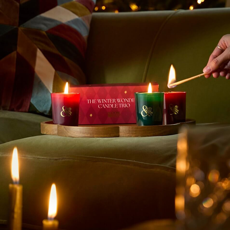 Three lit red and green Bloom & Wild candles in front of a Winter Wonder Candle Trio gift box on a wooden tray.