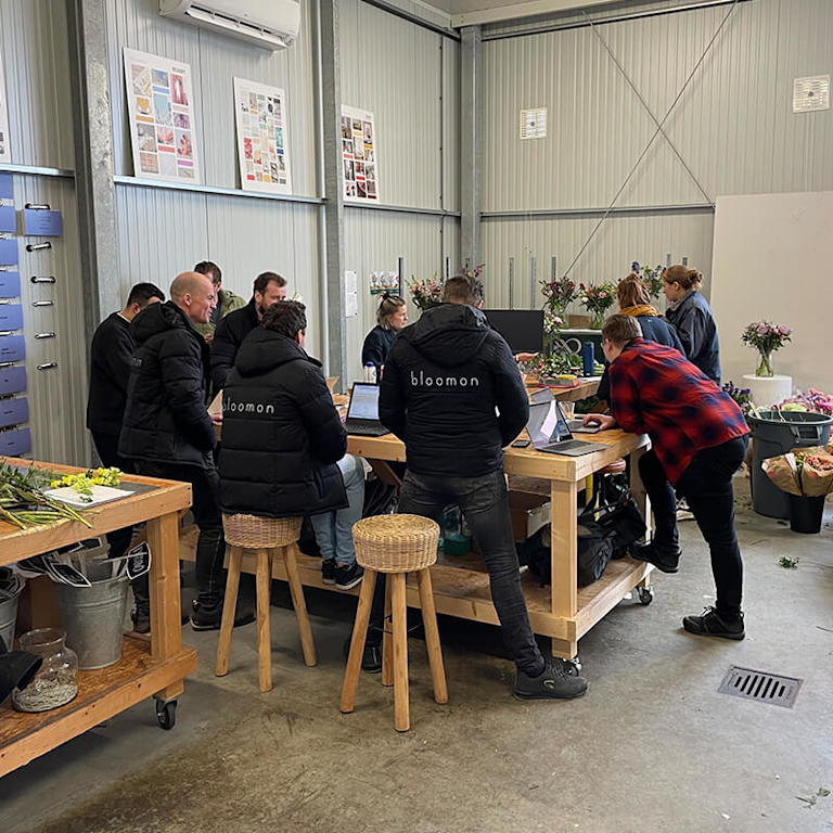 A group of people in bloomon hoodies gather around a high table to discuss floral designs. Several bouquets are lined up on plinths nearby.