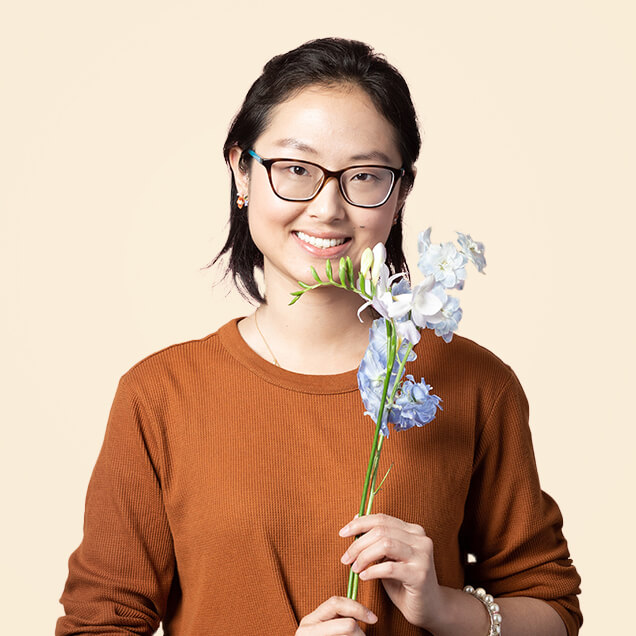Hanlin is a Chinese woman in her thirties. She wears glasses, and holds up a stem of a blue delphinium flower.