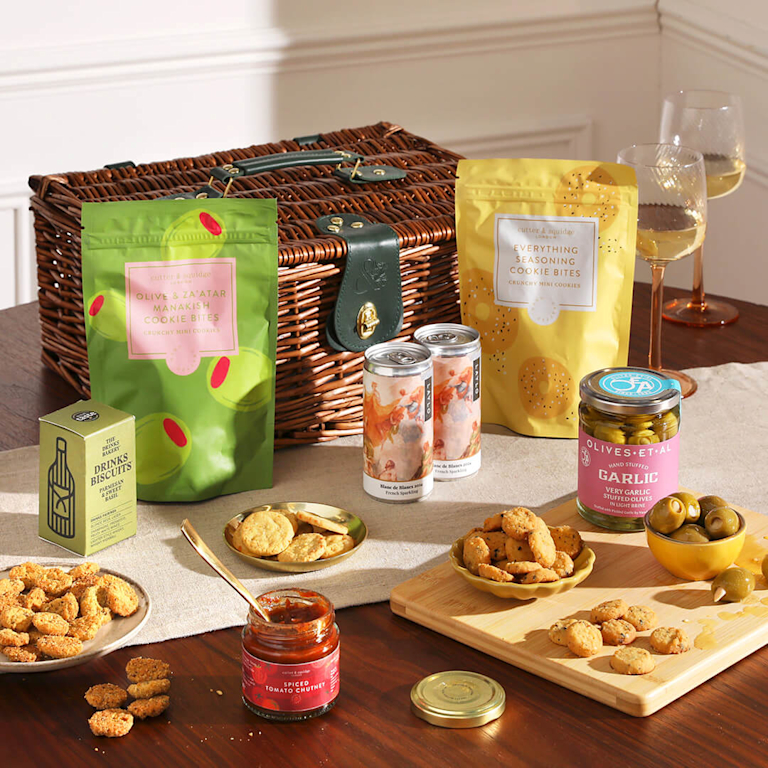 An abundant picnic hamper overflowing with savoury treats, including garlic-stuffed olives, spiced tomato chutney, and savoury biscuits set alongside sparkling wine.