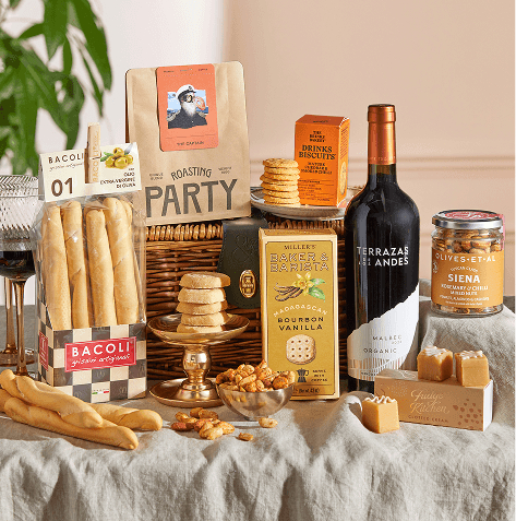 Wicker hamper with Malbec red wine, breadsticks, coffee, drinks biscuits, vanilla biscuits, a jar of mixed nuts, fudge and shortbread, arranged on a linen tablecloth.