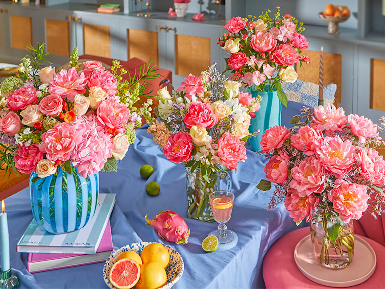 Bright kitchen scene with colorful floral bouquets (peonies, roses, more) in various vases on a light blue island. Champagne glasses, rosé, fruit, and decor add to the cheerful atmosphere.