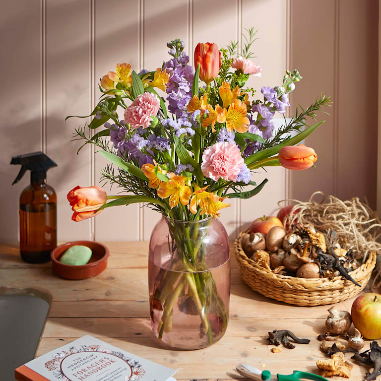 A rustic, meadow-style bouquet of lilac stocks, pink carnations and orange tulips in a mauve glass vase, set against a cosy, wooden kitchen backdrop.