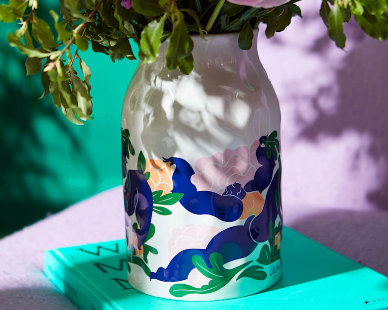 A close-up of a white ceramic vase with a Kelly Anna design on. The design features a bold, abstract female figure leaping through several large pink and peach peonies.