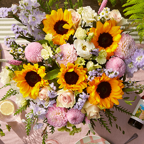 Overhead view of a bright bouquet with sunflowers, pink roses, dahlias, and delphinium on a table with a gingham cloth and lemon slice.