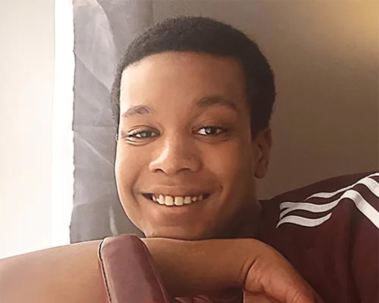 Alex, a dark-skinned young man in an Adidas t-shirt, smiles as he leans his arm on the back of a chair.