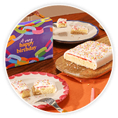 Colourful "It's your Happy Birthday" gift box beside a slice of pink-frosted sprinkle cake on a plate with a fork, next to the whole cake.
