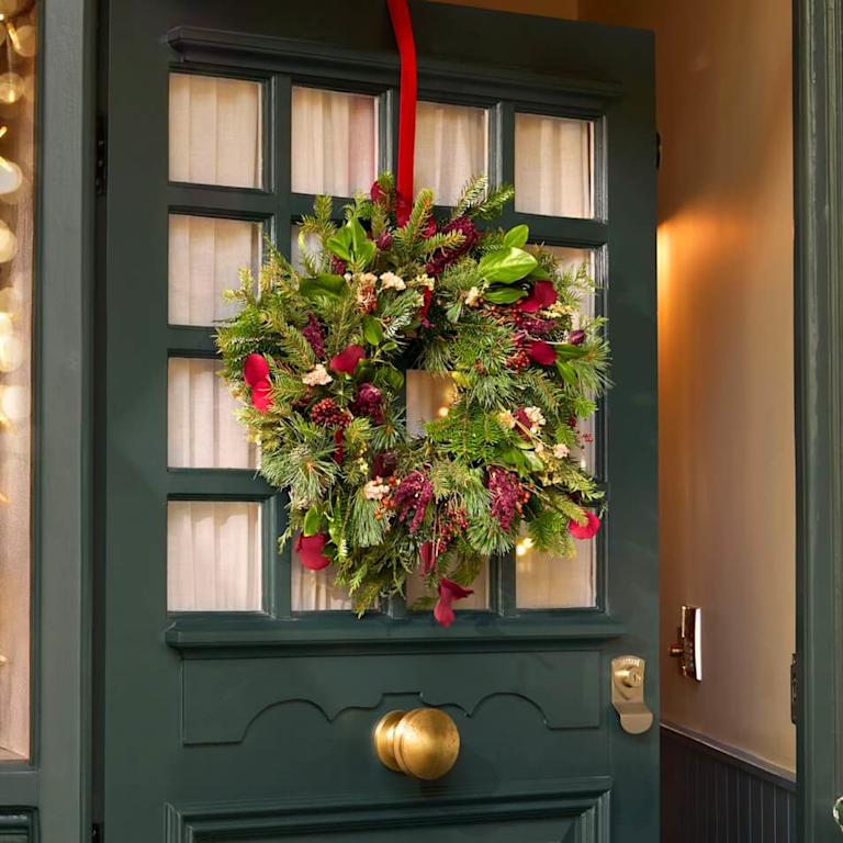 A lush Christmas wreath of mixed foliage, pine cones and deep red blooms hanging on a dark green front door.