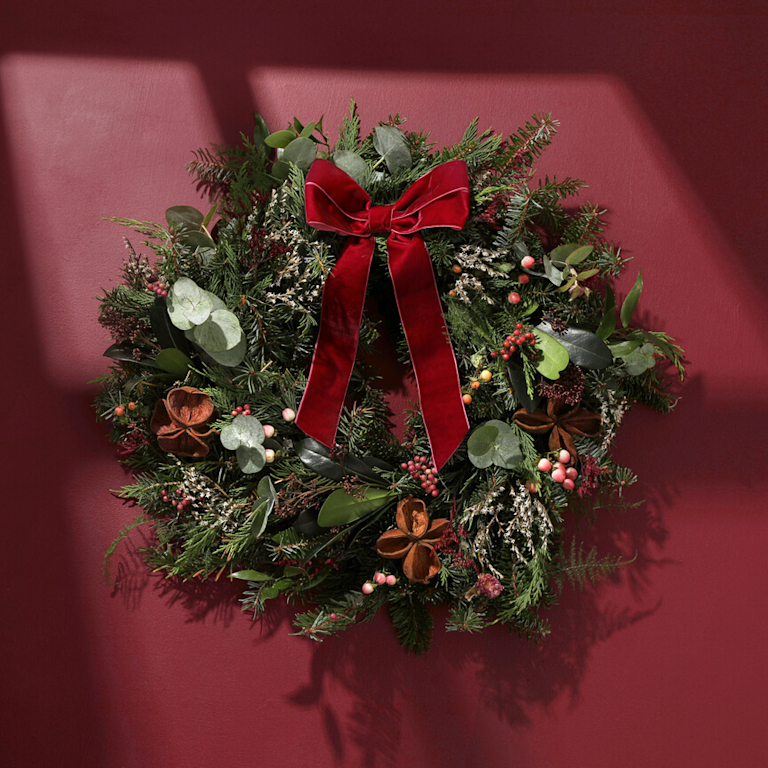A wreath, decorated with hypericum berries, dried stems and a red velvet ribbon against a red background.