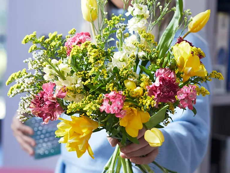 Two images side by side: the first shows a pretty bouquet in a green vase with yellow and white flowers; the second shows a person's hand unboxing a letterbox Bloom & Wild bouquet.