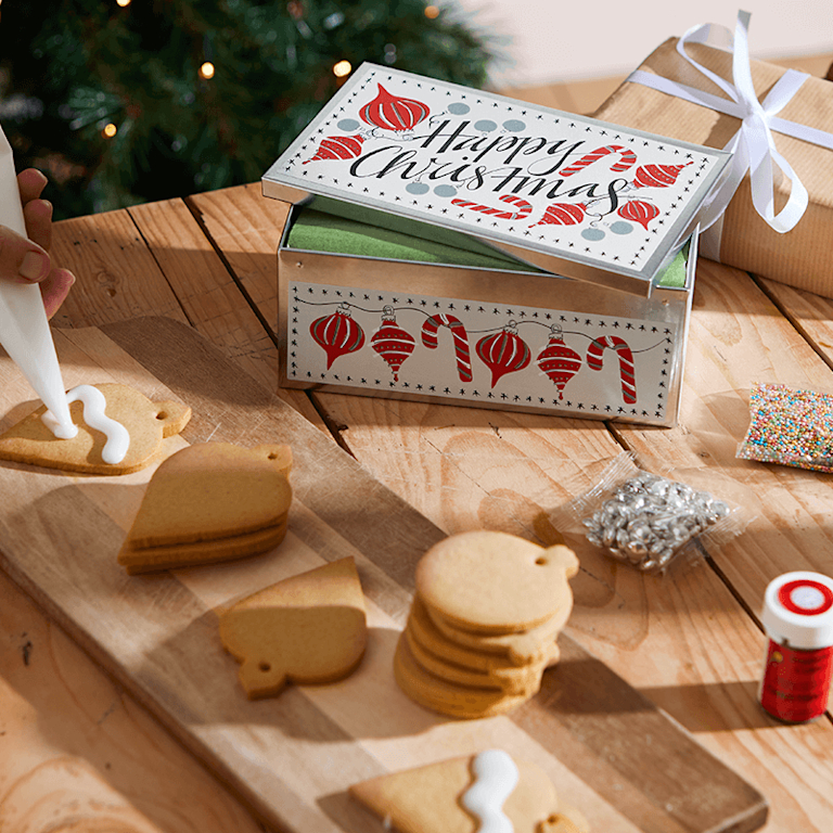 A biscuit tin decorated with the words 'Happy Christmas'. In front of it are bauble-shaped biscuits, along with icing and sparkly sprinkles.