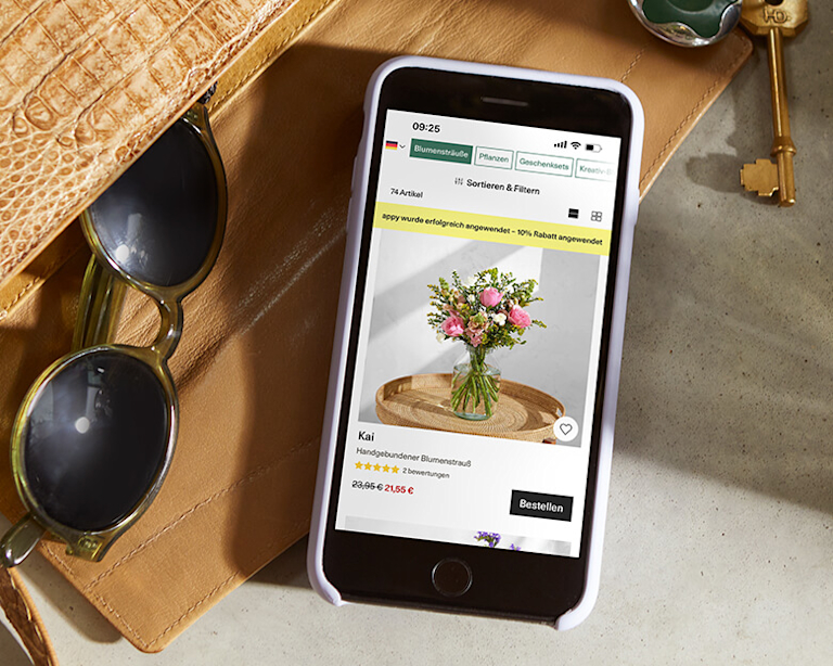A smartphone lays on top of an open purse. Other items, like sunglasses and keys, have spilled out beside it. The Bloom & Wild app appears on the smartphone screen.