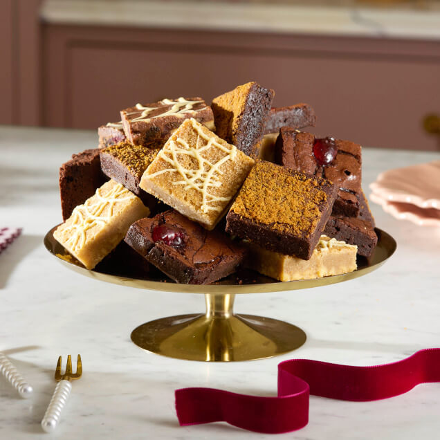 A gold cake stand piled high with chai-spiced blondies and chocolate brownies, with a red ribbon on a marble table.