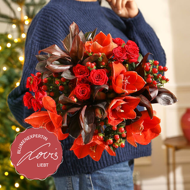 Festlicher Bouquet Lovis mit leuchtend roten Amaryllis und Rosen, kombiniert mit tiefrotem Laub und Beeren, gehalten vor einem Lichterbaum.