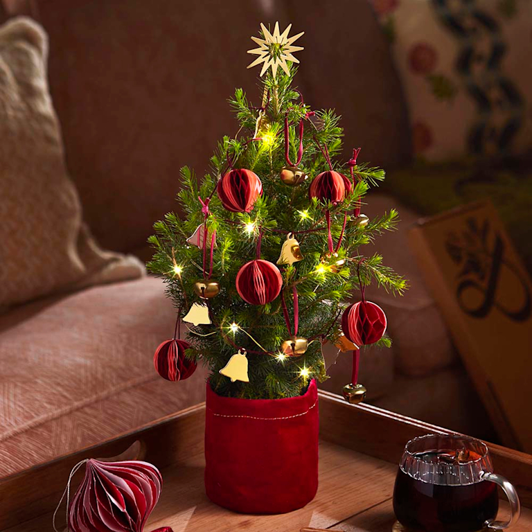 Kleiner, geschmückter Tannenbaum in einem roten Samttopf, dekoriert mit roten Papierkugeln, goldenen Glöckchen und Lichterkette, auf einem Holztablett mit Glühwein in gemütlicher Atmosphäre.
