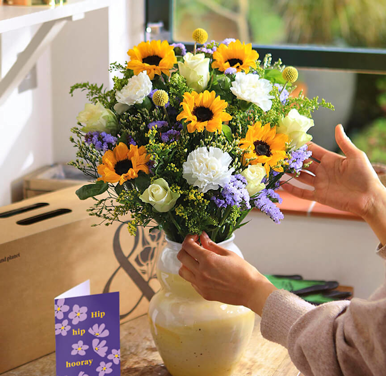 A pair of hands adjusts a bright bouquet of sunflowers, white carnations, cream roses, and purple statice in a cream vase. A 'Hip hip hooray' card and brown delivery box are nearby.